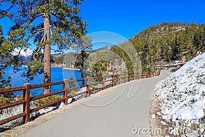 A gorgeous smooth concrete paved hiking trail with a burnt orange wooden fence along the trail with lush green pine trees and snow Stock Photo