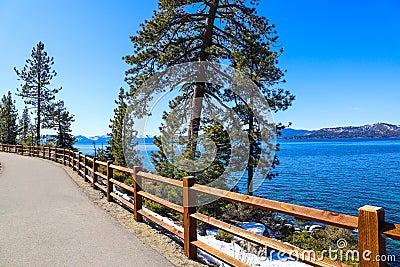 A gorgeous smooth concrete paved hiking trail with a burnt orange wooden fence along the trail with lush green pine trees and snow Stock Photo