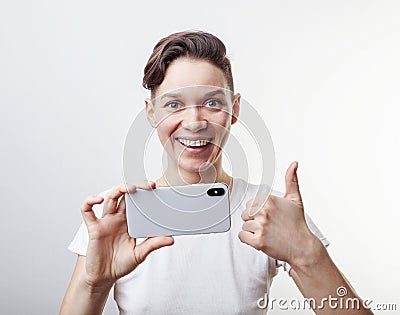 Gorgeous teenage girl is taking a photo using front camera in her smartphone and showing thumb up. Isolated on white background Stock Photo