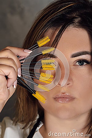 Gorgeous young lashmaker holding yellow disposable eyelash extension brushes close to face standing on gray background. Stock Photo