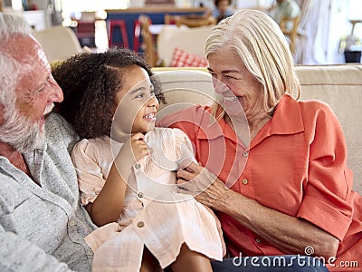 Granddaughter Cuddling With Loving Grandparents On Sofa At Home With Parents In Background Stock Photo