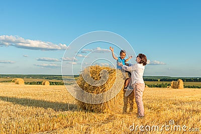 A grandmother with a grandson puts on a haystack on a field on a sunny day. Grandson delighted, they have fun together Stock Photo