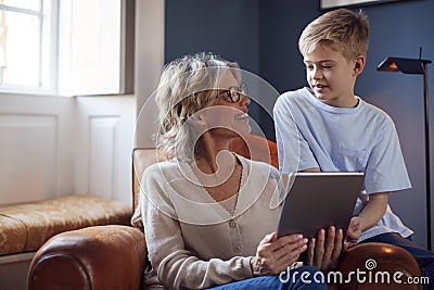 Grandson With Grandmother Sitting In Chair Playing On Digital Tablet At Home Together Stock Photo