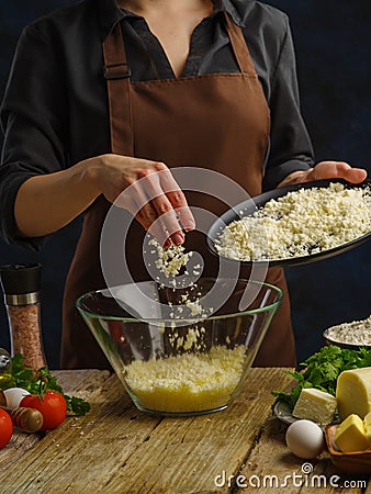 Grated cheese in a large glass bowl in frozen flight on a dark background. Vegetables, greens, eggs, seasonings on a wooden table Stock Photo