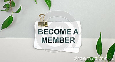 On a gray background - green leaves of the plant and a white card under a gold clip with the text BECOME A MEMBER. Minimalist Stock Photo