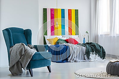 Gray blanket on a comfy, blue armchair next to a colorful bed with cushions in a bright bedroom interior with striped painting on Stock Photo