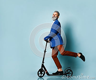 Gray-haired elderly man in white shirt, jacket, brown pants, loafers. Riding black scooter and posing sideways on blue background Stock Photo