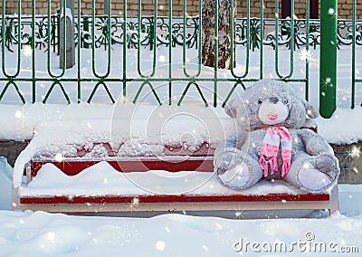 A gray plush toy hare with a pink scarf sits on a bench in the snow in winter, forgotten by a child. The theme of sadness, loneli Stock Photo