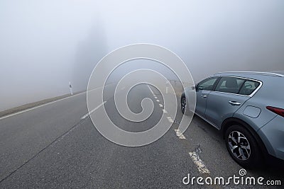 A gray SUV car is parked on the side of a straight empty road invaded by thick fog. In the background, the silhouettes Stock Photo