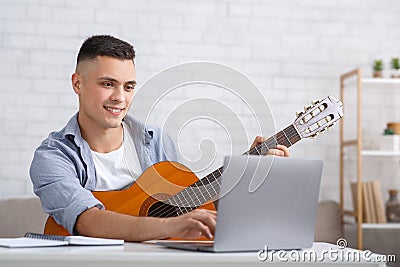 Great mood and new hobby during coronavirus epidemic in free time at home. Cheerful guy with guitar works in laptop and Stock Photo
