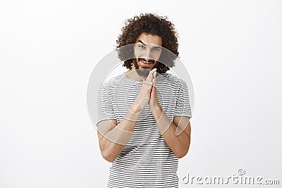 Greedy guy has nasty plan. Portrait of intrigued curly-haired man with beard, rubbing palms together near chest and Stock Photo