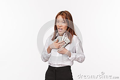 Greedy young rich asian female in white shirt, press money to chest and squinting suspicious, scheming, stole cash Stock Photo