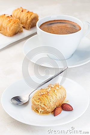 Greek pastry Kataifi with shredded filo dough stuffed with almond nuts, in honey syrup, on a white plate, served with cup of Stock Photo