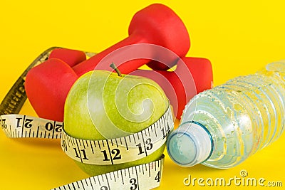 Green apple with measuring tape, red dumbbell and fresh water bottle with drops on yellow background Stock Photo