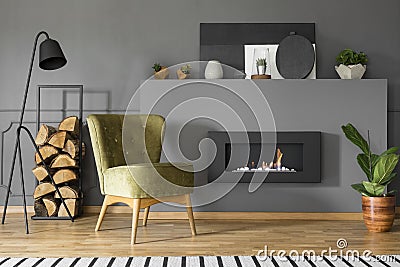 Green armchair and firewood next to fireplace in grey living room interior with lamp. Real photo Stock Photo