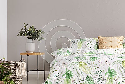 Green flower in grey vase on wooden bedside table next to comfortable bed with floral bedding, copy space on the empty grey wall Stock Photo