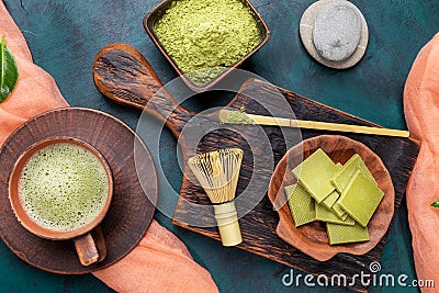 Green matcha tea and powder in brown ceramic cups and matcha chocolate on wooden serving board on emerald background. Stock Photo