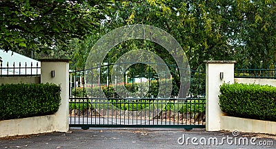 Green metal wrought iron driveway property entrance gates set in concrete fence, garden shrubs, trees, lights Stock Photo