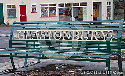 A green park bench in the town centre of Glastonbury. The name of the town is featured in large gold lettering Editorial Stock Photo