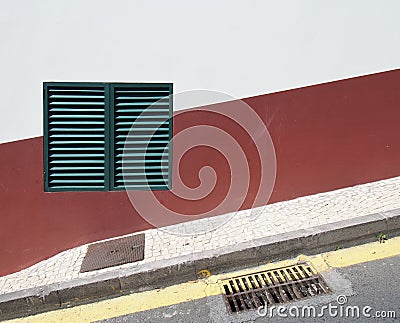 a green shuttered closed window on a red and white house wall on a sloping street with yellow parking line and pavement in typical Stock Photo