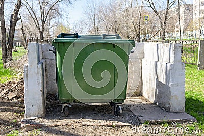 A green trash can stands on a site with a white concrete fence in a public park. Garbage collection and disposal using a plastic Stock Photo