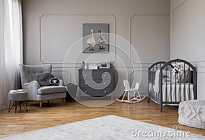 Grey chest of drawers in the middle of elegant grey baby room with comfortable armchair and wooden cradle Stock Photo