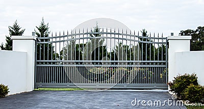 Grey metal wrought iron driveway property entrance gates set in white concrete brick fence, garden trees in background Stock Photo