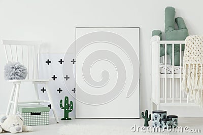 Grey pompom placed on white chair standing in bright baby bedroom interior with cactus shaped decor and empty poster on the floor Stock Photo