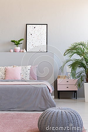 Grey pouf next to bed with cushions in modern bedroom interior with poster and plants. Real photo Stock Photo