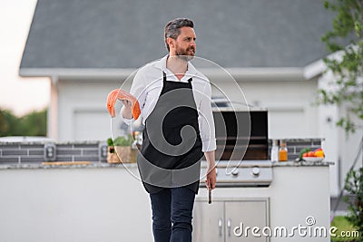 Grill cook. Chef in cook apron with BBQ cooking tools. Barbecue and grill. Chief cook hold salmon fish with utensils for Stock Photo