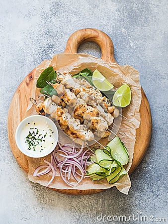 Grilled chicken on metal skewers on spinach leaves, slices cucumber, red onion and lime and sauce in a bowl on wood board Stock Photo