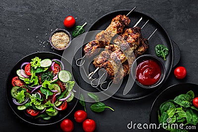 Grilled meat skewers, shish kebab and healthy vegetable salad of fresh tomato, cucumber, onion, spinach, lettuce and sesame on bla Stock Photo