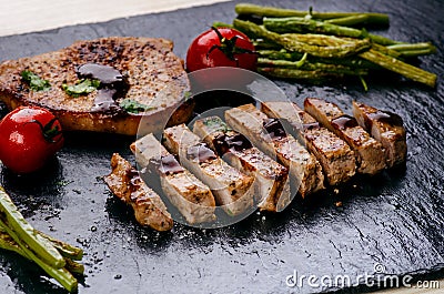 Grilled pork chops, steaks with vegetables, tomatoes, beans and sauce on a black slate. Fresh meat with foam. Dark background. Gri Stock Photo