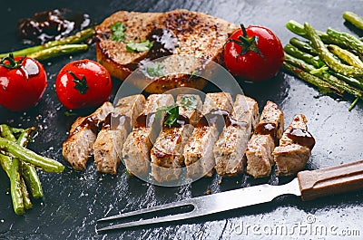 Grilled pork chops, steaks with vegetables, tomatoes, beans and sauce on a black slate. Fresh meat with foam. Dark background. Gri Stock Photo