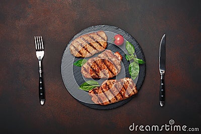 Grilled pork steaks with basil, tomatoes, knife and fork on black stone and brown rusty background Stock Photo