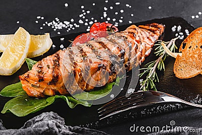 Grilled salmon fillet garnished with spinach, lemon, herbs on on stone board on black table surface. Hot fish dish Stock Photo