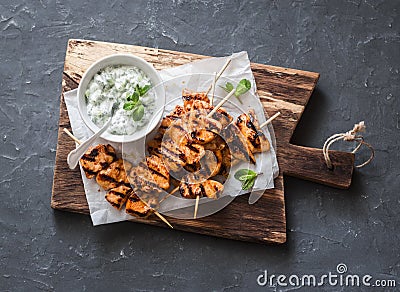 Grilled teriyaki chicken skewers and tzatziki sauce on a wooden board on a dark background, top view. Stock Photo