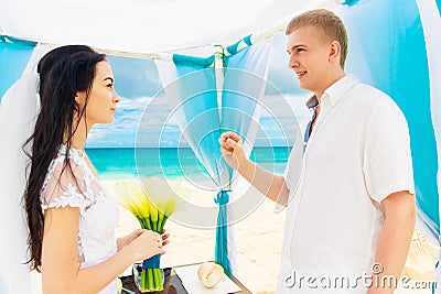 Groom giving an engagement ring to his bride under the arch decorated with flowers on the sandy beach. Wedding ceremony on Stock Photo