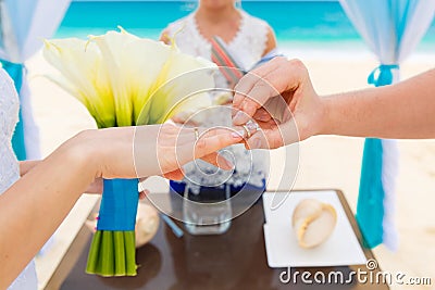 Groom giving an engagement ring to his bride under the arch decorated with flowers on the sandy beach. Wedding ceremony on Stock Photo