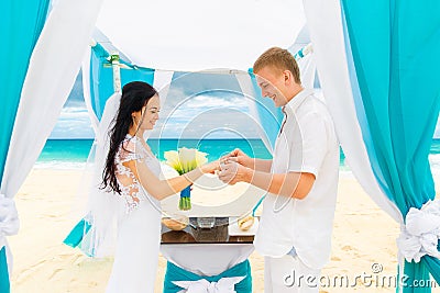 Groom giving an engagement ring to his bride under the arch decorated with flowers on the sandy beach. Wedding ceremony on Stock Photo