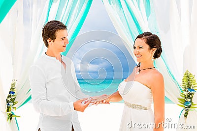 Groom giving an engagement ring to his bride under the arch decorated with flowers on the sandy beach. Wedding ceremony on Stock Photo