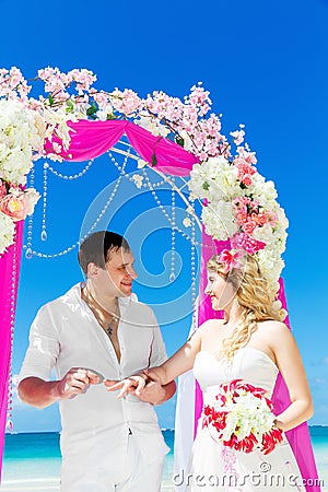 Groom giving an engagement ring to his bride under the arch decorated with flowers on the sandy beach. Wedding ceremony on Stock Photo