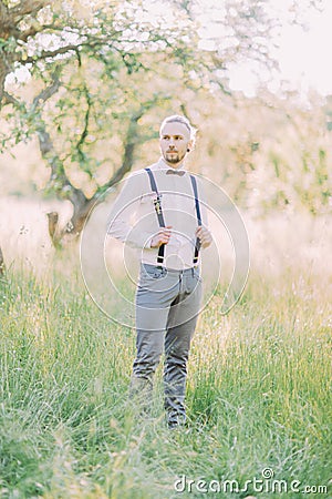 The groom in the gray pants and white shirt holding his black suspenders in the green spring field. Stock Photo