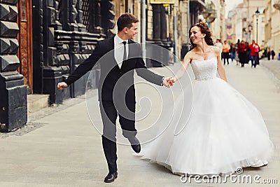 Groom holds bride`s hand tightly while they run along old buildi Stock Photo
