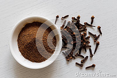 Ground cloves in white ceramic bowl isolated on white wood background from above. Whole cloves. Stock Photo