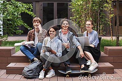 Group of cool smiling students sitting and happily looking in camera while spending time together in courtyard of Stock Photo