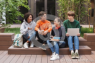 Group of cool smiling students sitting and happily looking at each other while spending time together in courtyard of Stock Photo