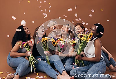 Group of diverse women with bouquets having fun under falling petals. Six happy females sitting together against a brown Stock Photo