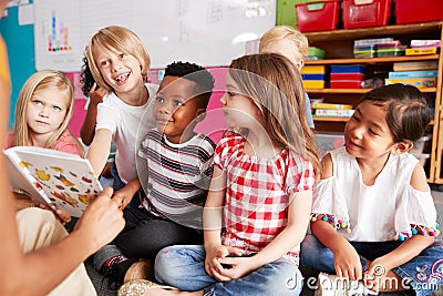 Group Of Elementary School Pupils Sitting On Floor Listening To Female Teacher Read Story Stock Photo