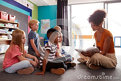 Group Of Elementary School Pupils Sitting On Floor Listening To Female Teacher Read Story Stock Photo